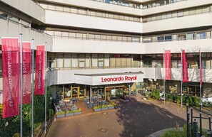 Een gebouw met de naam "Leonardo Royal" in grote letters. Er zijn meerdere rode vlaggen met tekst in het Cyrillisch alfabet, wat suggereert dat de locatie zich in een Russischtalig gebied bevindt. Het gebouw heeft een moderne architectuur met grote ramen en een plat dak.