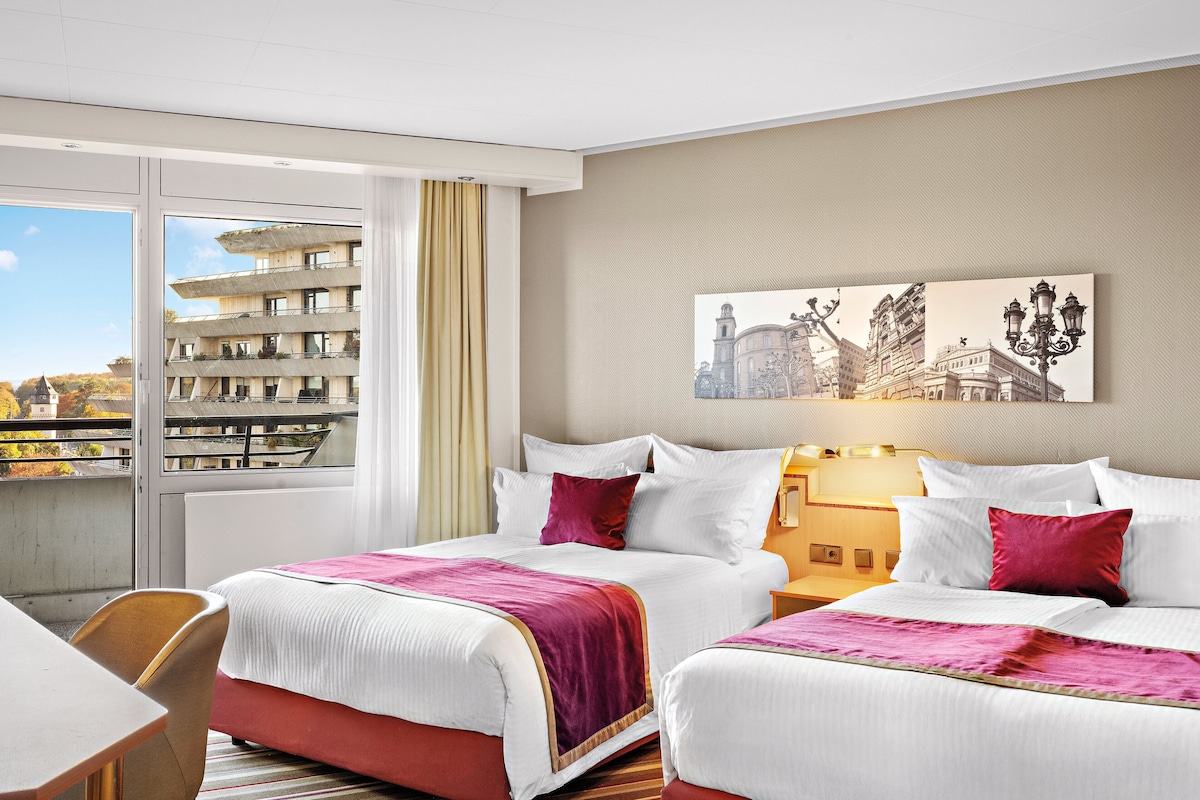 A hotel room with two beds, a large window with a view of a building, a painting on the wall, and a nightstand with a lamp.