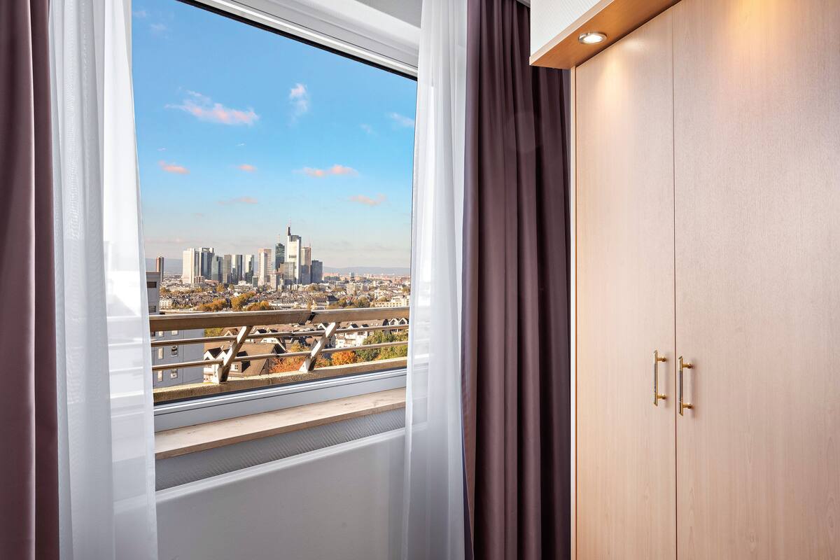Une chambre dotée d’une grande fenêtre donnant sur le paysage urbain, d’une armoire en bois et d’un balcon avec une balustrade.