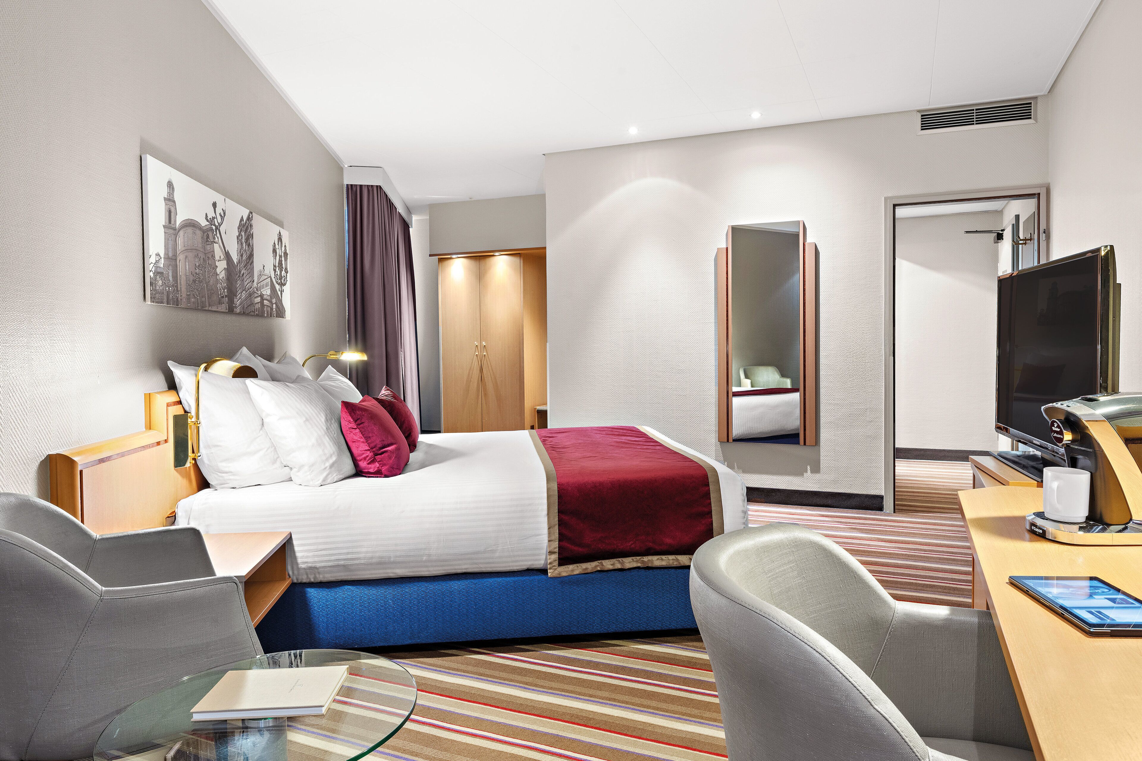 A hotel room with a bed, a desk with a TV, a chair, a glass table, and a large mirror.