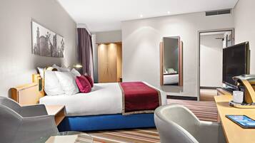 A hotel room with a bed, a desk with a TV, a chair, a glass table, and a large mirror.