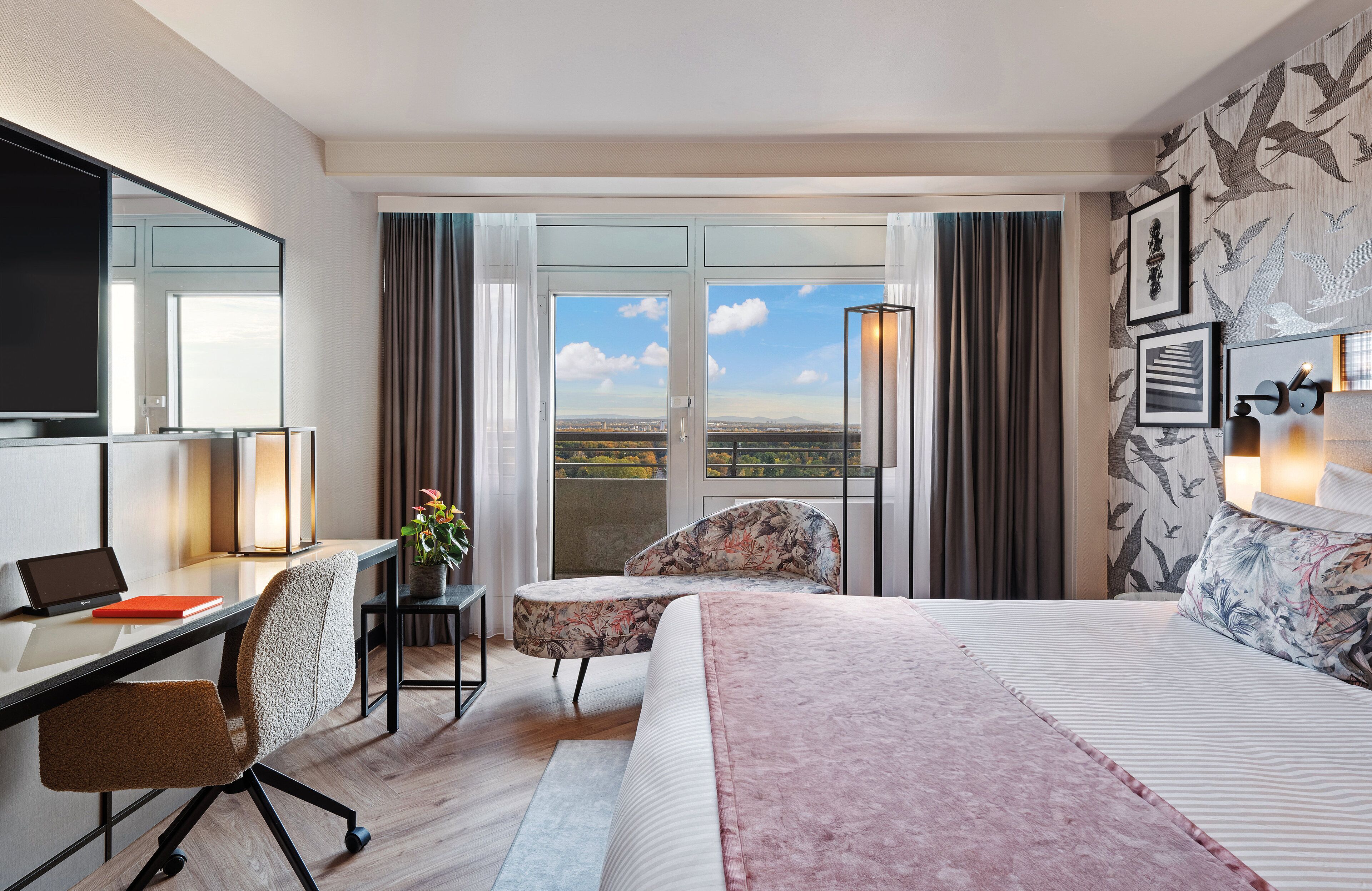 Ein modernes Hotelzimmer mit einem großen Bett, einem Schreibtisch mit einem Laptop, einem Stuhl und Blick auf die Stadt durch große Fenster.