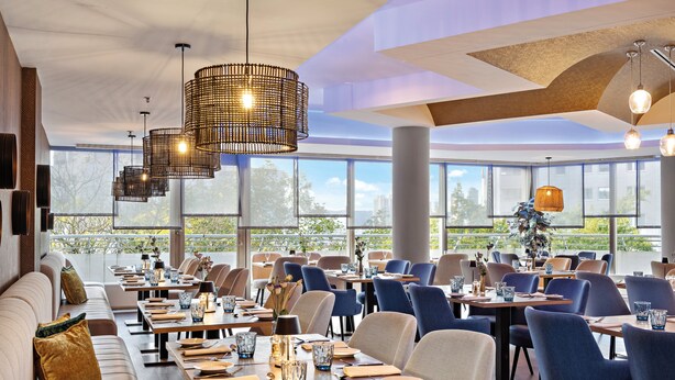 Een modern restaurant met hoge plafonds, grote ramen en diverse zithoekopstellingen, waaronder tafels en stoelen.