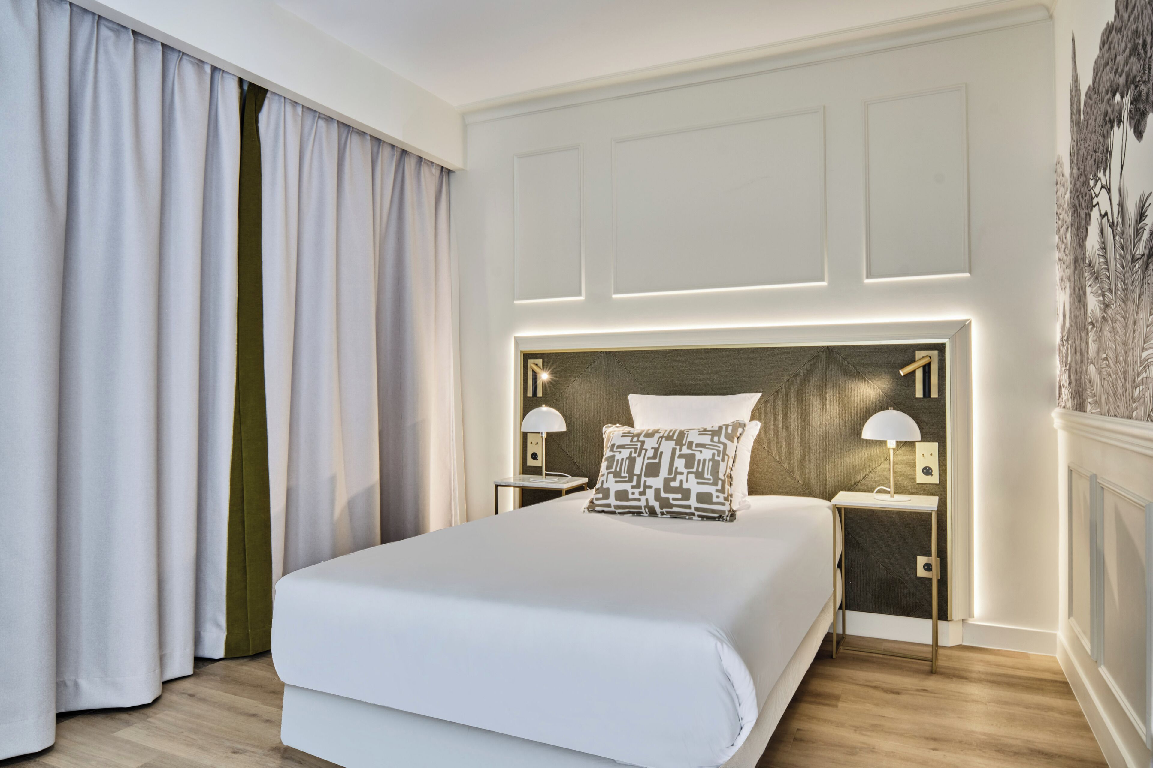 Une chambre d’hôtel dotée d’un grand lit, de deux tables de chevet avec des lampes, d’une tête de lit avec un éclairage intégré et d’une vue sur l’extérieur à travers des rideaux.