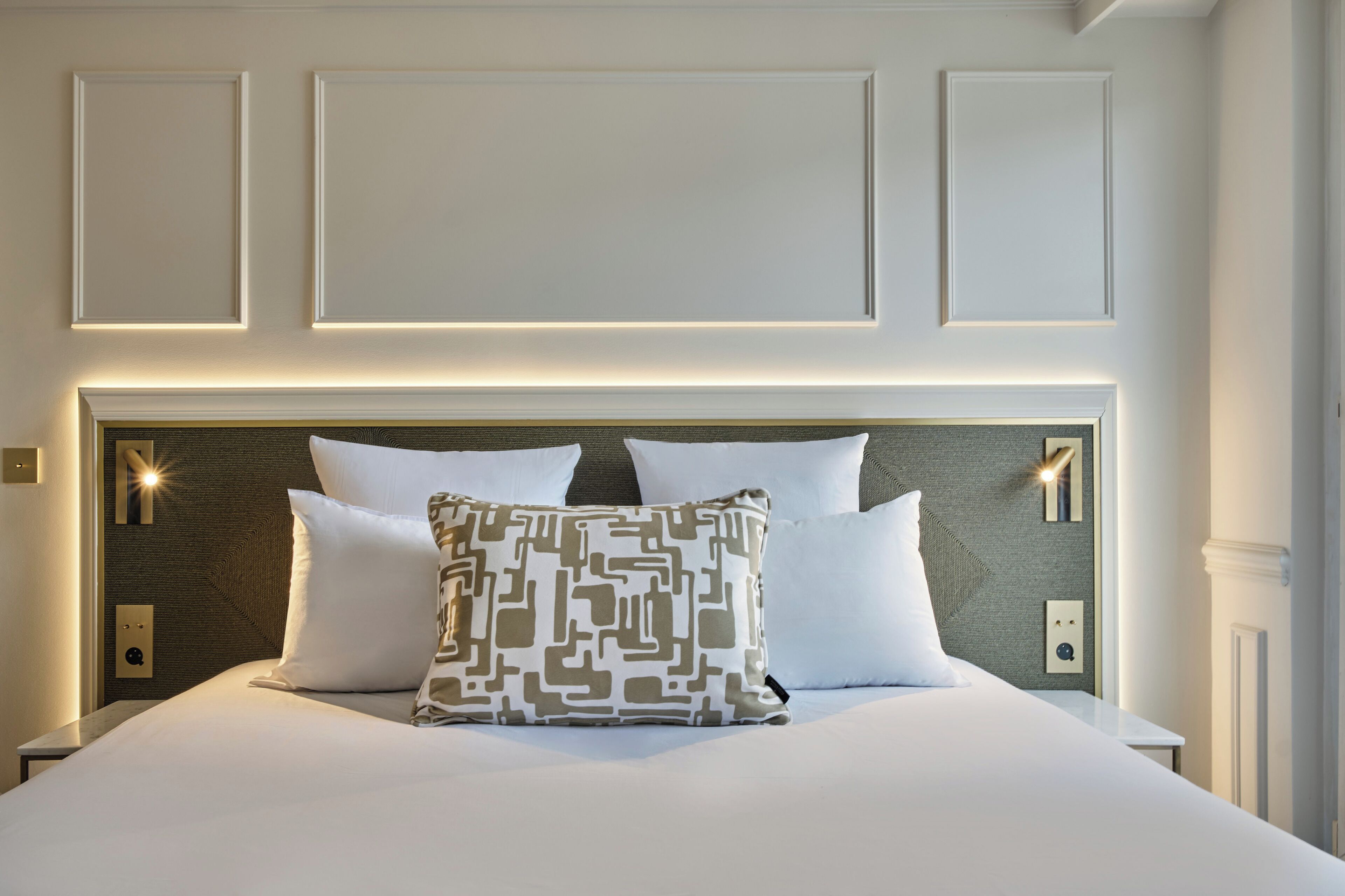 Una cama bien tendida con una almohada estampada, cabecera con iluminación integrada y pared con paneles empotrados.