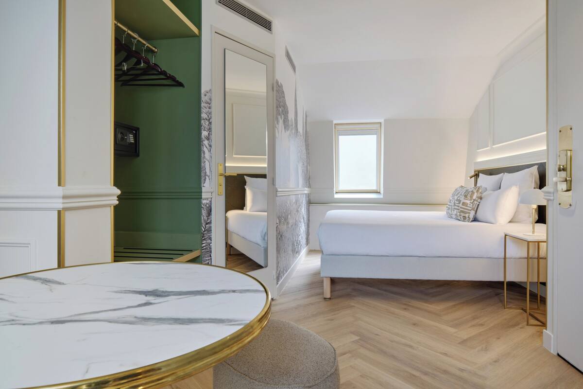Camera d'albergo moderna con un tavolo in marmo, un letto con biancheria bianca, un piccolo comodino e una finestra che offre luce naturale.