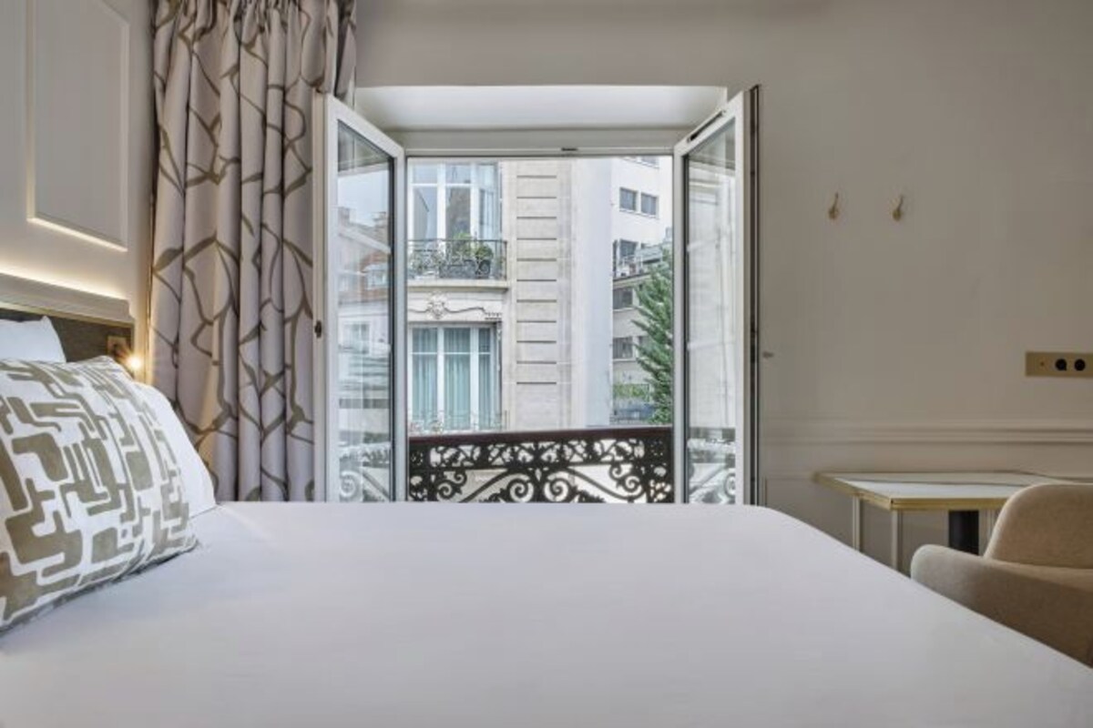 A hotel room with a bed, a chair, a small table, and a balcony with a railing.