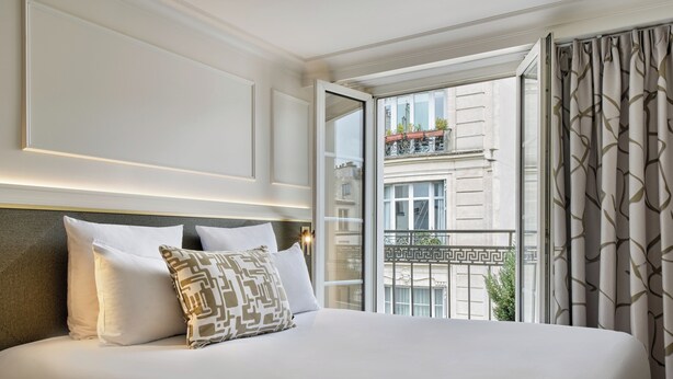 A hotel room with a bed, a nightstand, and a window with a view of a building and balcony. - Posy Hotel by Happyculture (Paris)