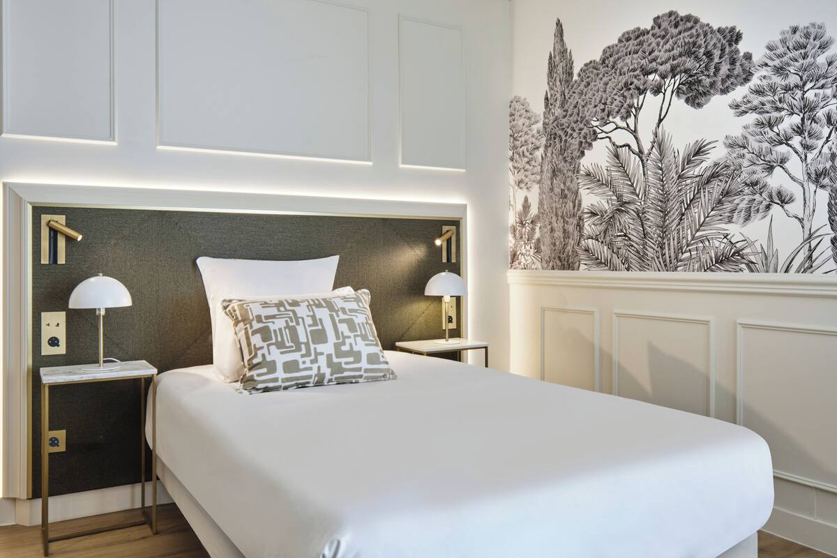 A neatly made bed with a white duvet and patterned pillows, flanked by two bedside tables with lamps, in a room with a detailed wall mural and a modern headboard.
