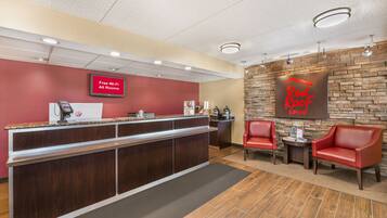 A hotel reception area with a red wall, a 'Free Wi-Fi All Rooms' sign, a 'Red Roof Inn' sign, and two red leather chairs.