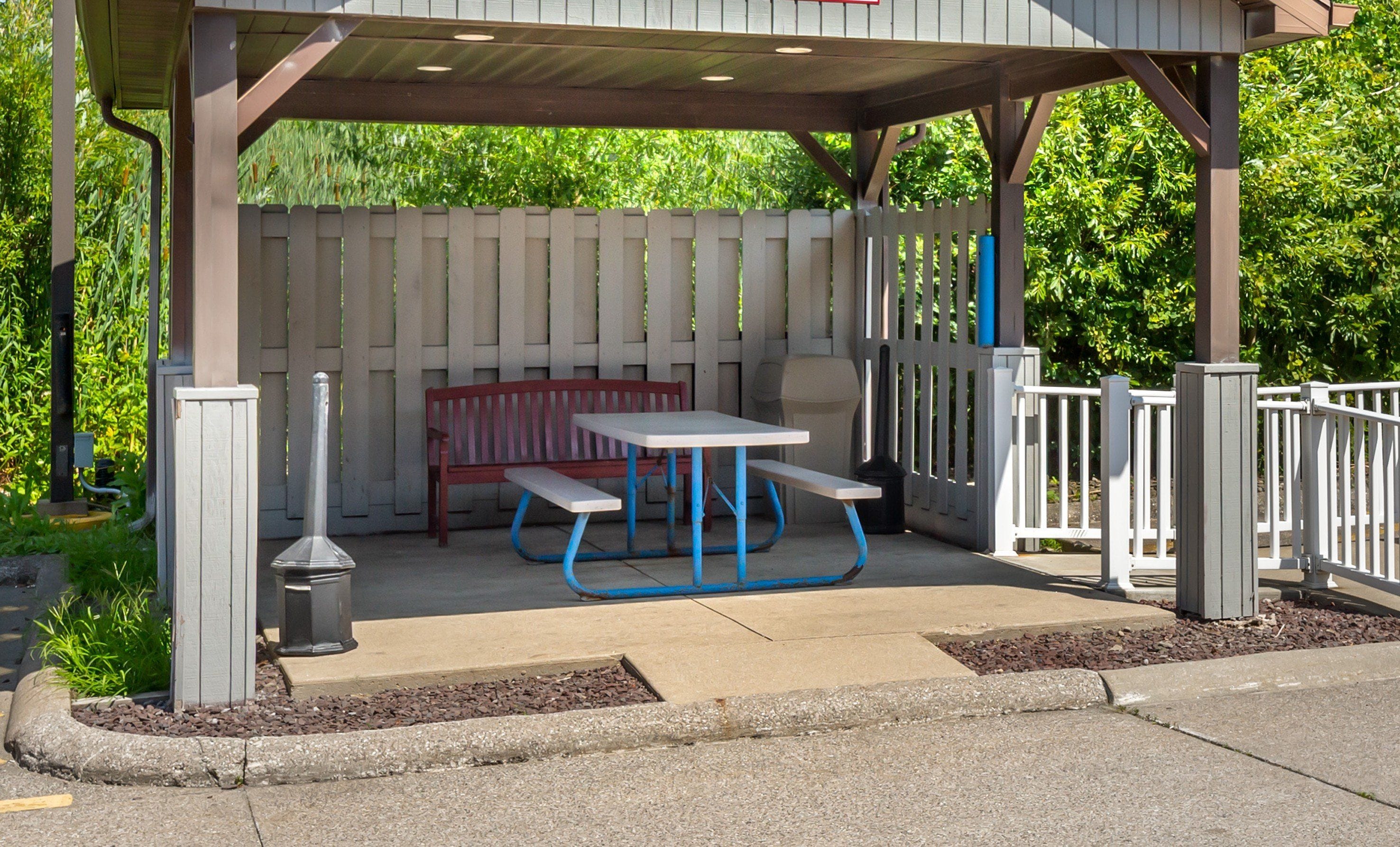 A covered outdoor seating area with a picnic table and benches, surrounded by a white fence and greenery.