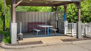 A covered outdoor seating area with a picnic table and benches, surrounded by a white fence and greenery.