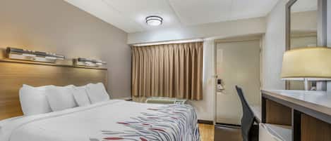 A hotel room with a bed, a desk, a chair, a desk lamp, a window with curtains, and a door.