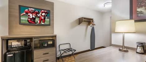 Habitación de hotel con televisión de pantalla plana en la pared, minibar, escritorio con lámpara y una silla.