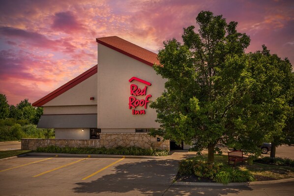 Un hotel Red Roof Inn con fachada de piedra, un estacionamiento y un camino de entrada bordeado de árboles.
