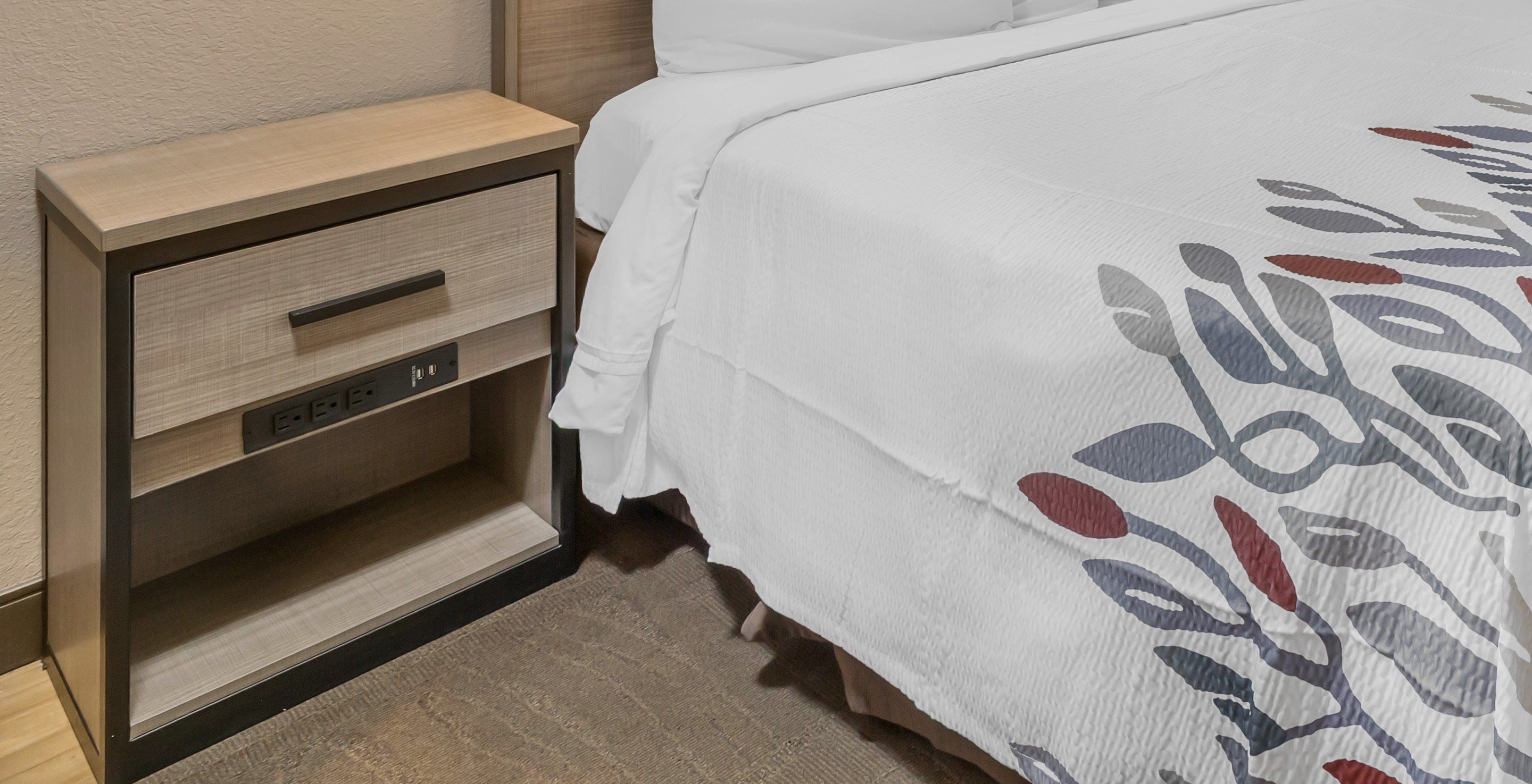 A neatly made bed with a white bedspread featuring a leaf pattern, a wooden nightstand with a drawer, and a power outlet.