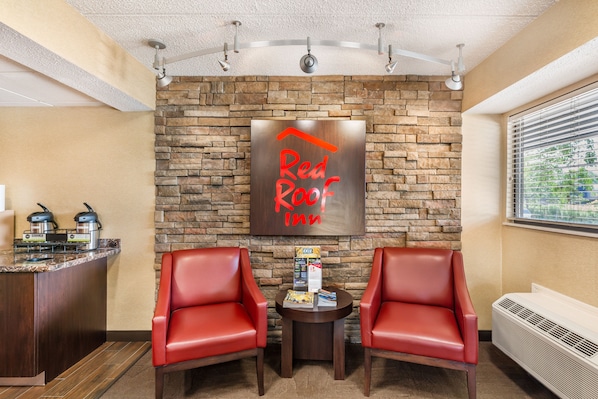 A Red Roof Inn lobby with a stone wall, two red leather chairs, a small table with amenities, and a view through the window. - Red Roof Inn Erie – I-90 (Erie)