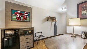 A hotel room with a wooden desk, a black refrigerator, a TV mounted on the wall, a lamp, and a coat rack. - Red Roof Inn Erie – I-90 (Erie)