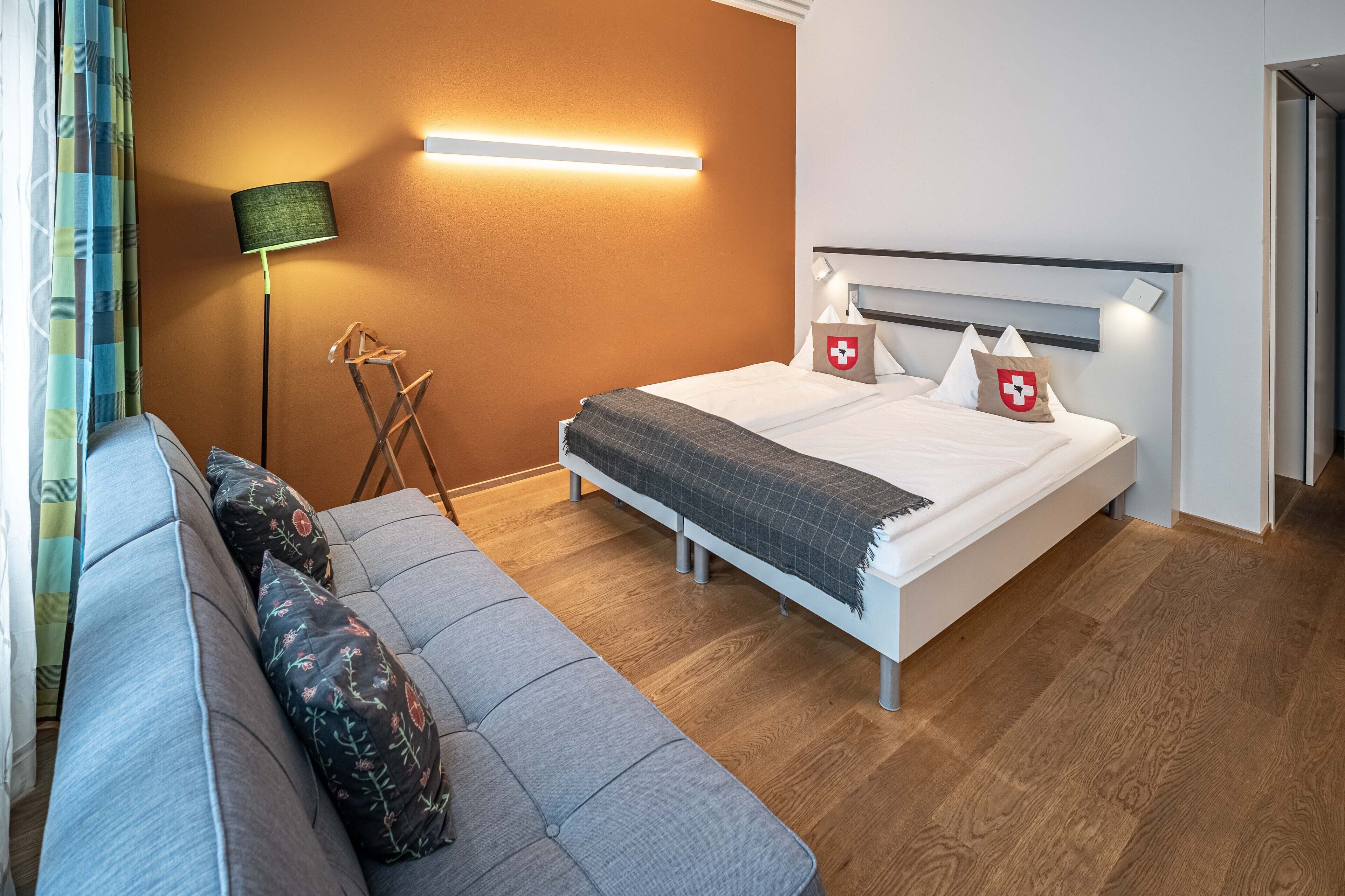 A modern hotel room with a bed, a sofa, a standing lamp, and a wall-mounted light.