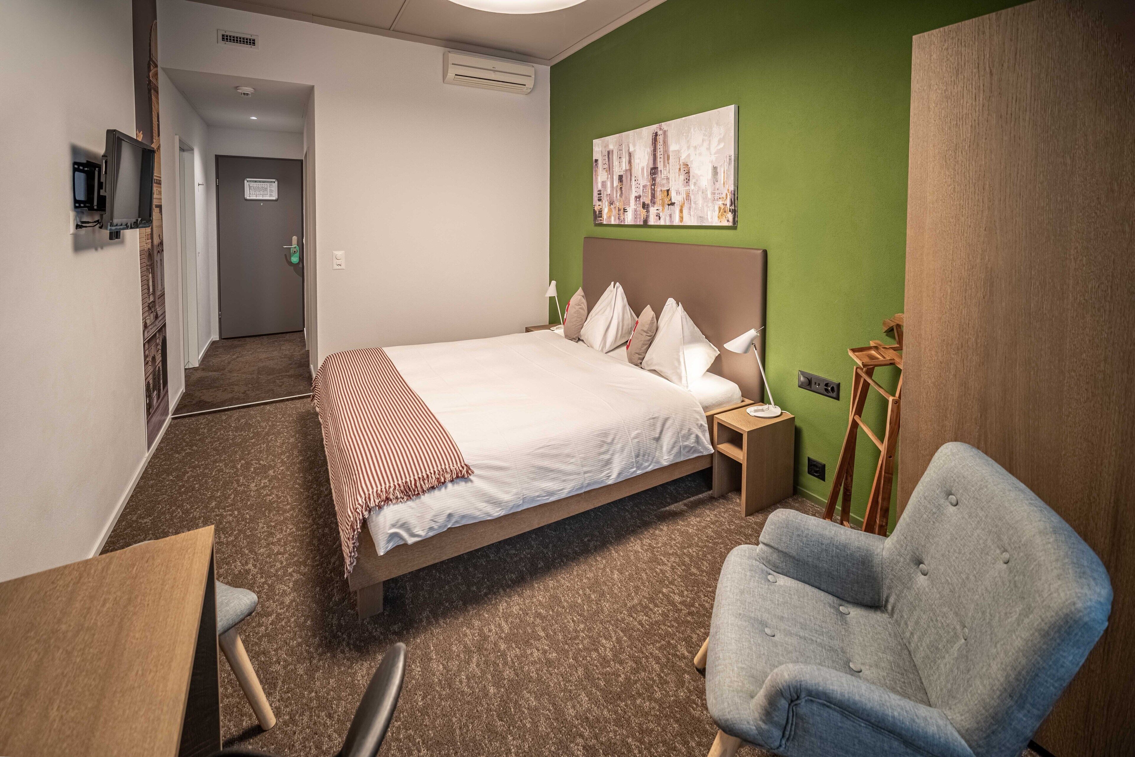 Un cuarto de hotel con una cama grande, un sillón gris, una mesita y un cuadro en la pared.