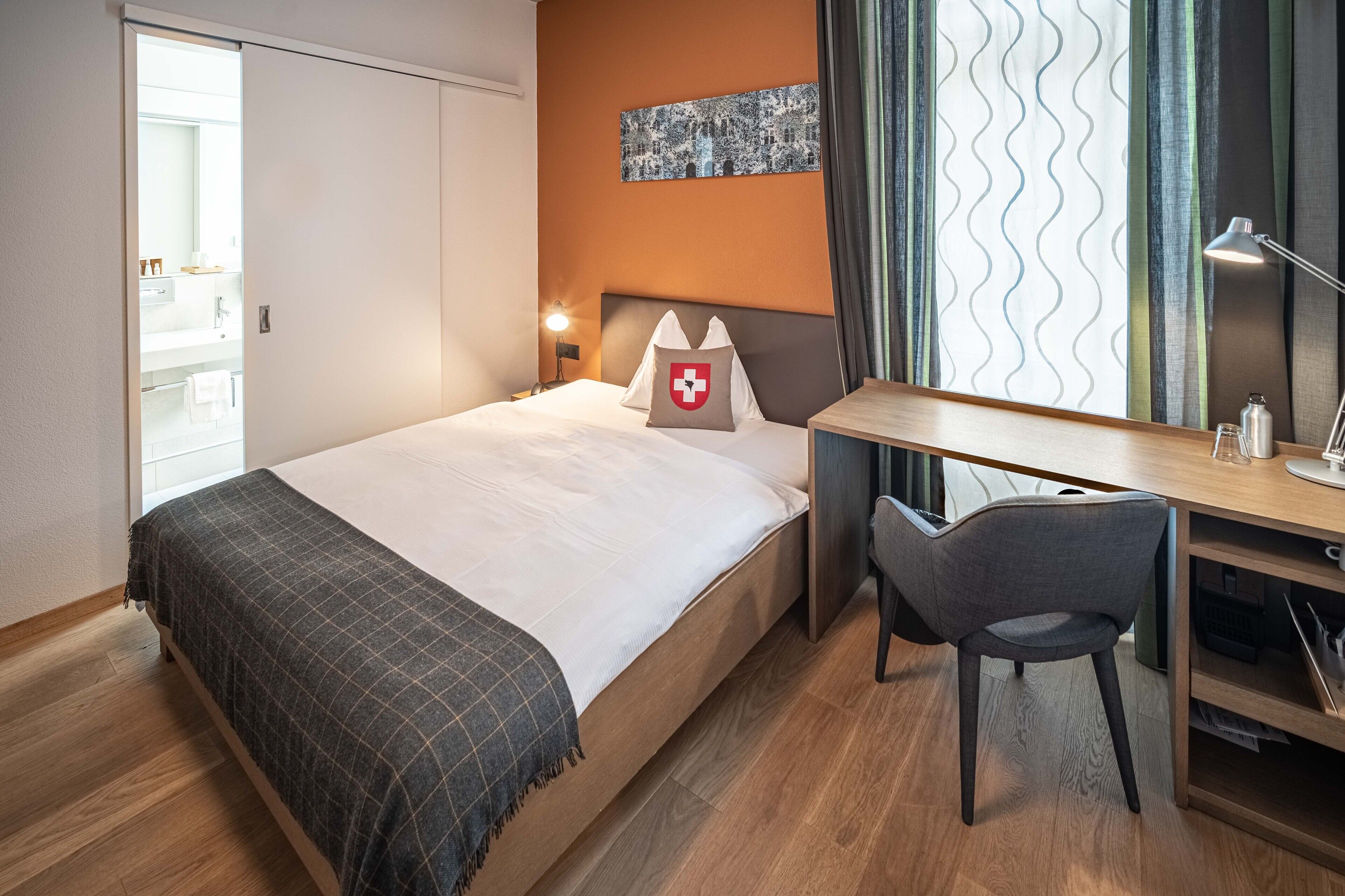 Habitación de hotel con cama, escritorio, silla, mesita de noche, lámpara y un cuadro en la pared.