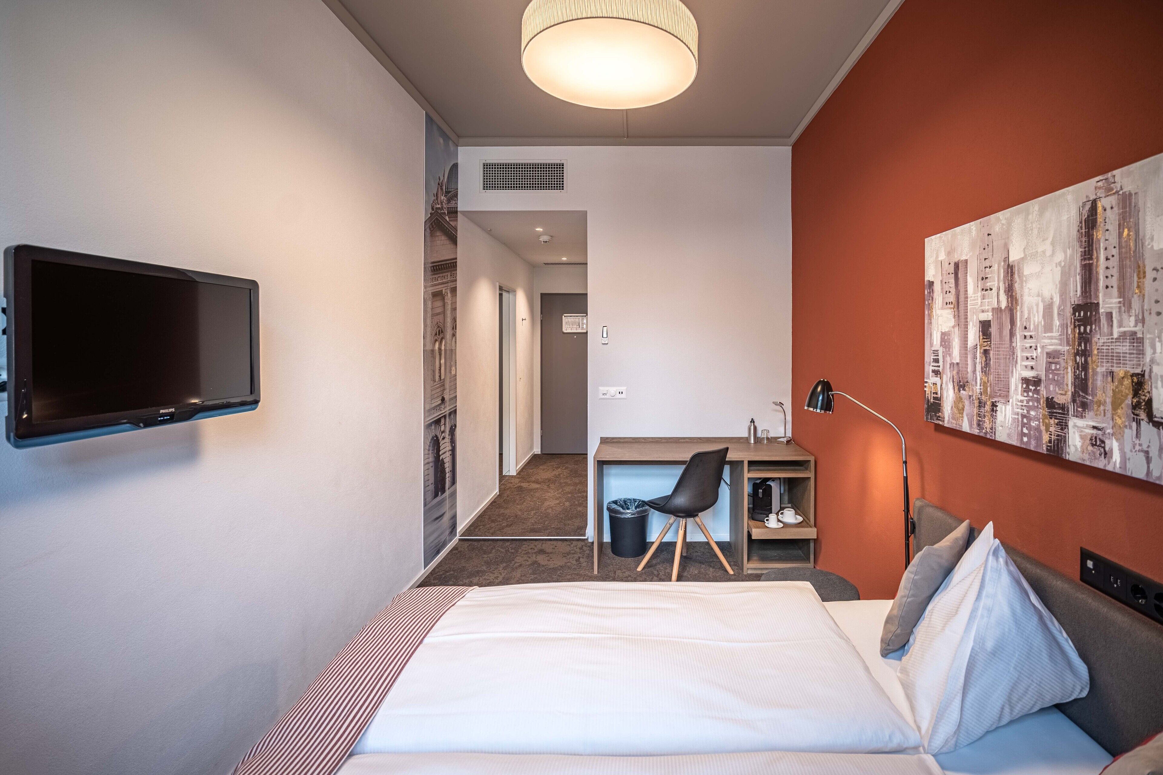Ein Hotelzimmer mit einem Bett, einem Schreibtisch mit Stuhl, einem an der Wand montierten Fernseher und einem Stadtbild an der Wand.