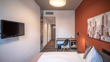 Ein Hotelzimmer mit einem Bett, einem Schreibtisch mit Stuhl, einem an der Wand montierten Fernseher und einem Stadtbild an der Wand.