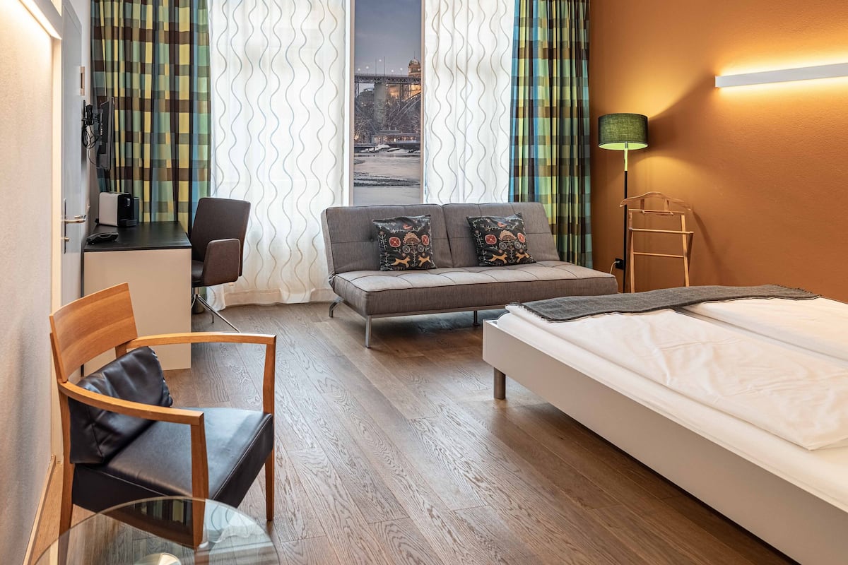 A hotel room with a bed, a sofa, a desk with a chair, a glass table, a lamp, and curtains.