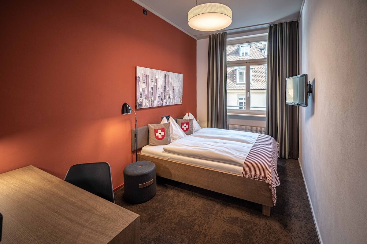 A hotel room with a bed, a desk, a chair, a small ottoman, a painting on the wall, a window with curtains, and a television mounted on the wall.