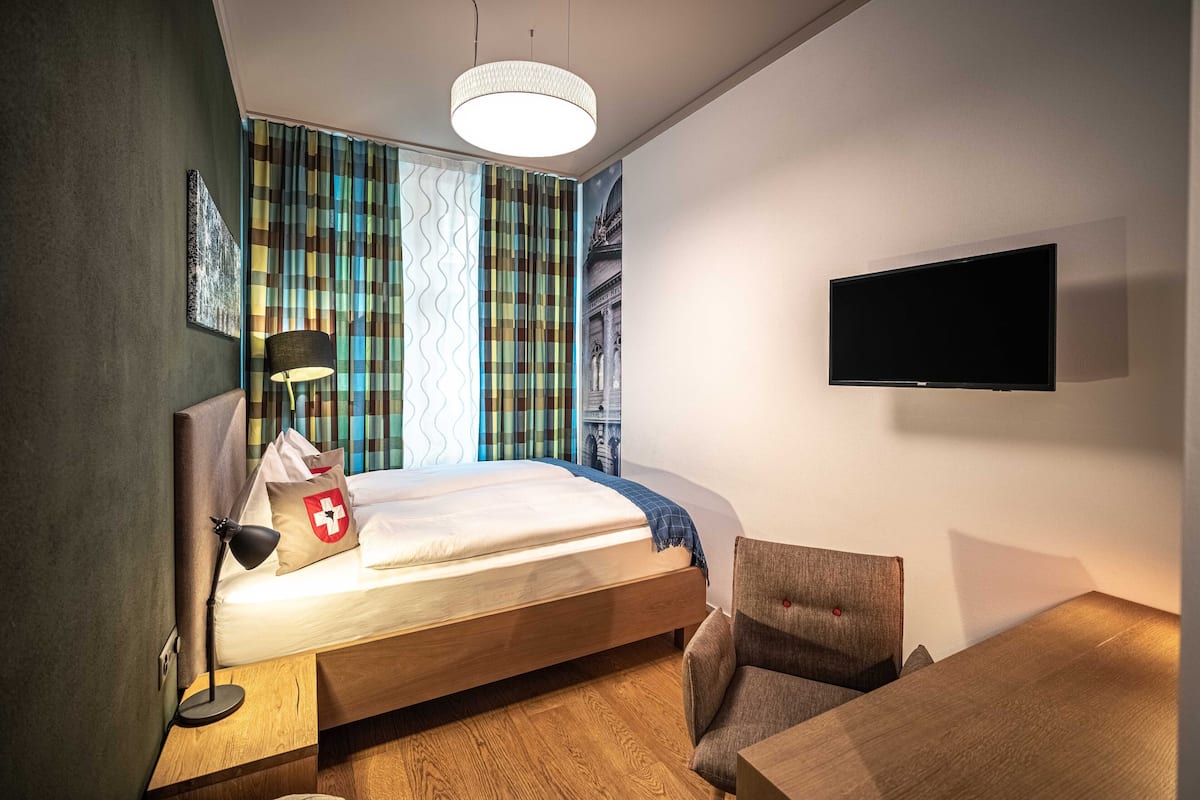A modern hotel room with a bed, a desk, a chair, a flat-screen TV, and a large window with curtains.