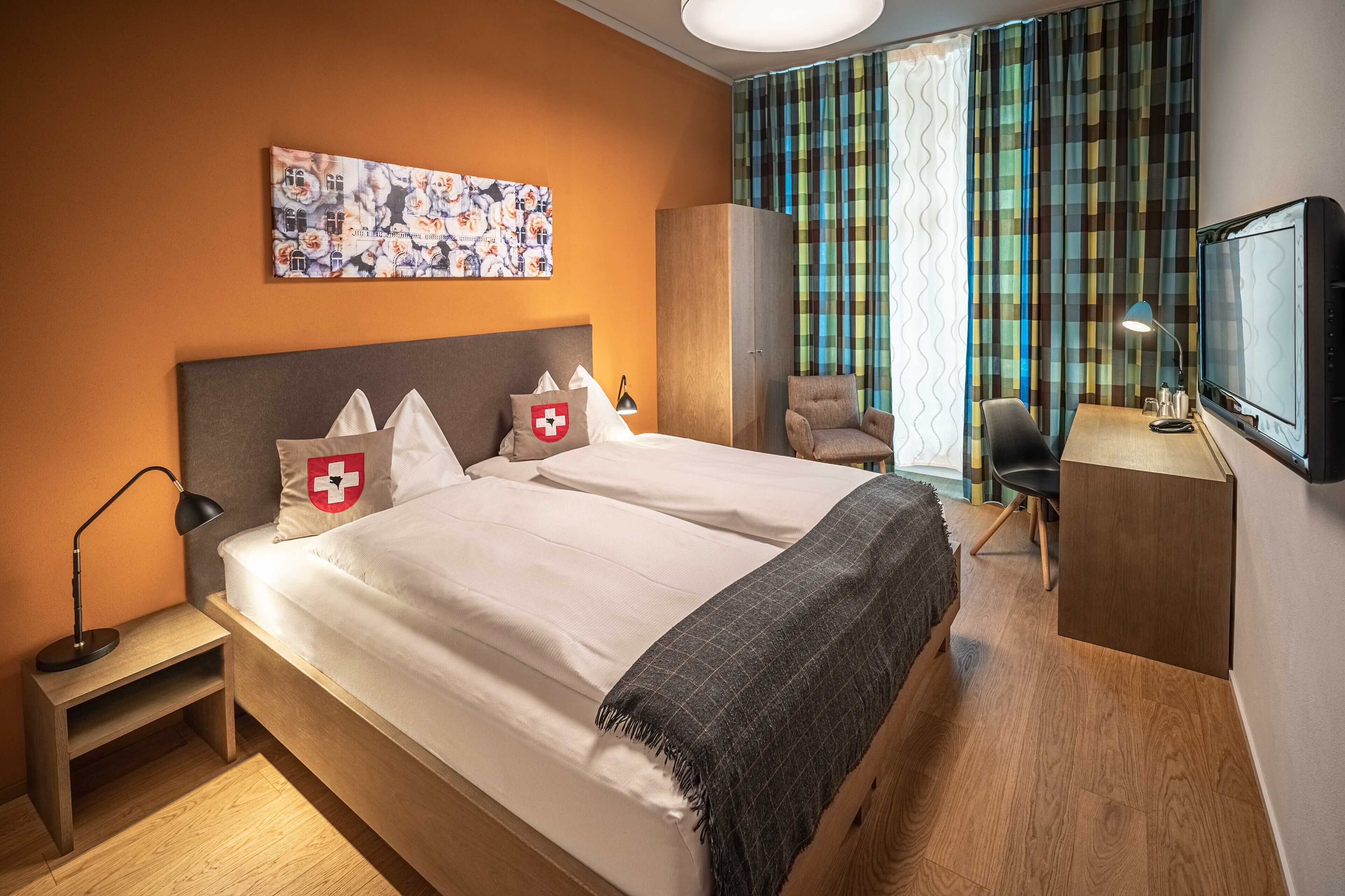 A hotel room with a large bed, a desk with a chair, a television, and a window with curtains.