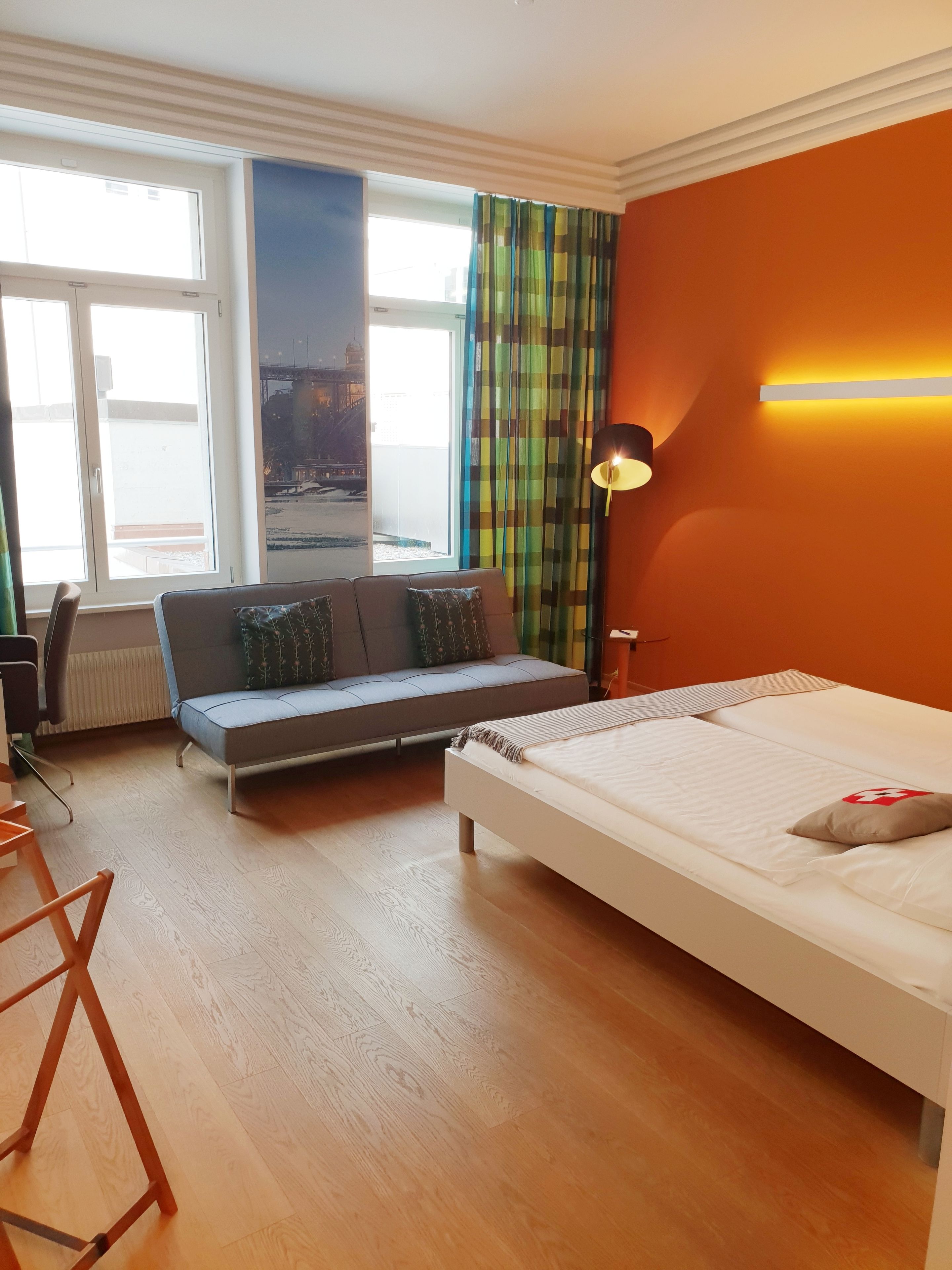 A modern hotel room with a bed, a sofa, a desk, and a view of the cityscape through large windows.
