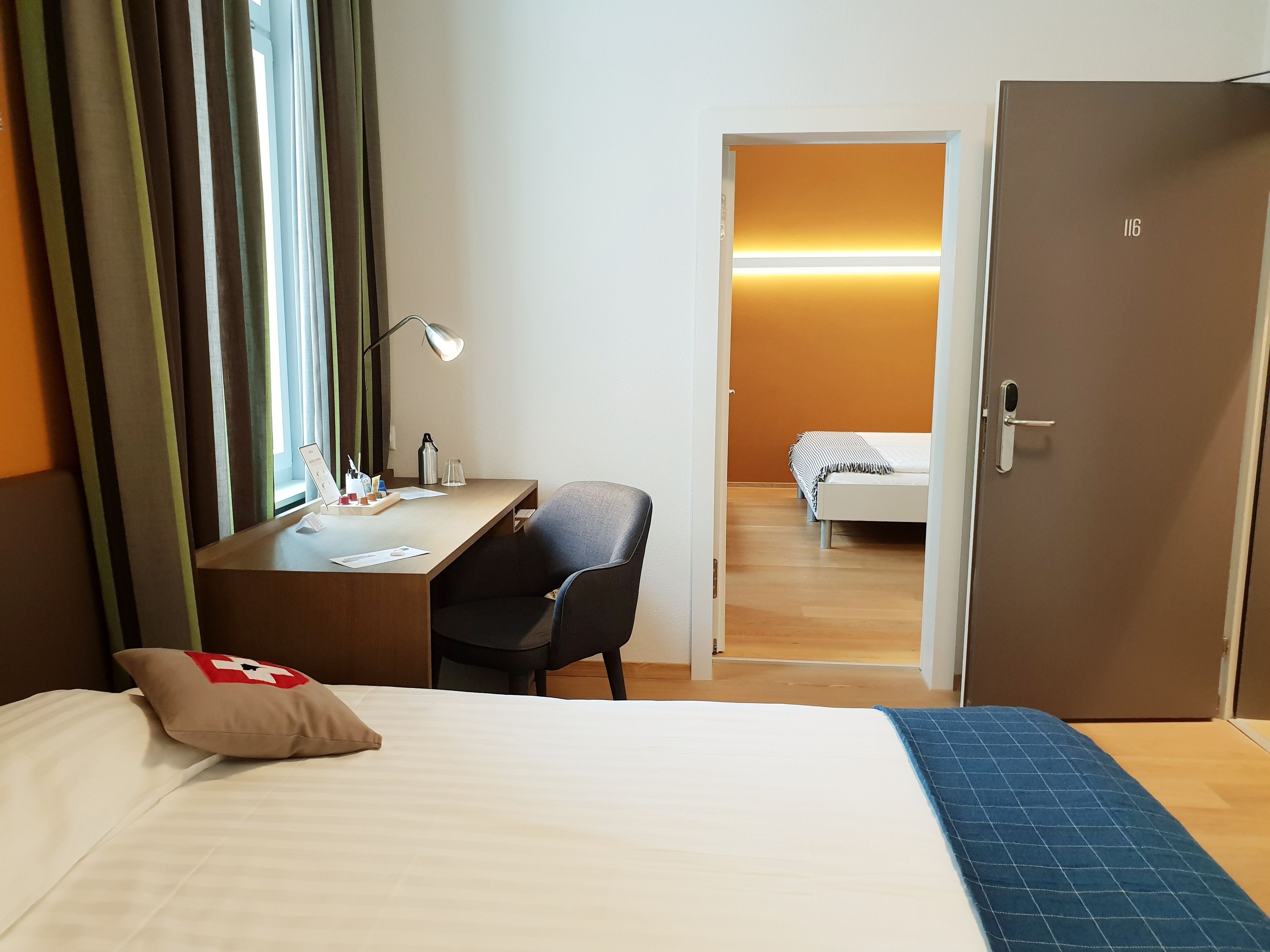 Quarto de hotel com cama, escrivaninha, cadeira e porta para outro cômodo.