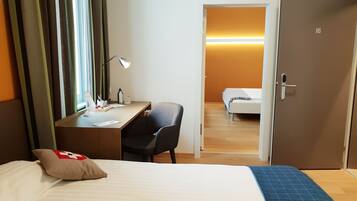 Quarto de hotel com cama, escrivaninha, cadeira e porta para outro cômodo.