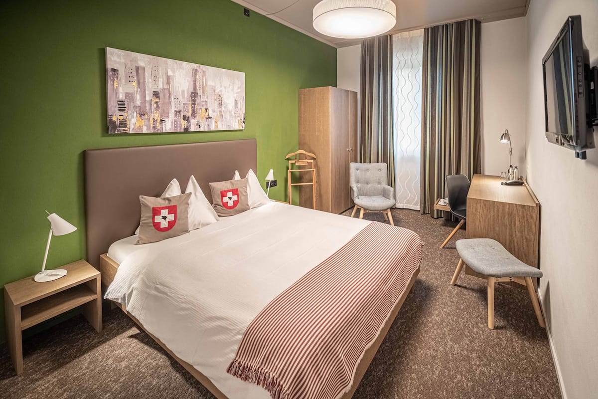A hotel room with a bed, a desk, a chair, a TV, and a painting on the wall.