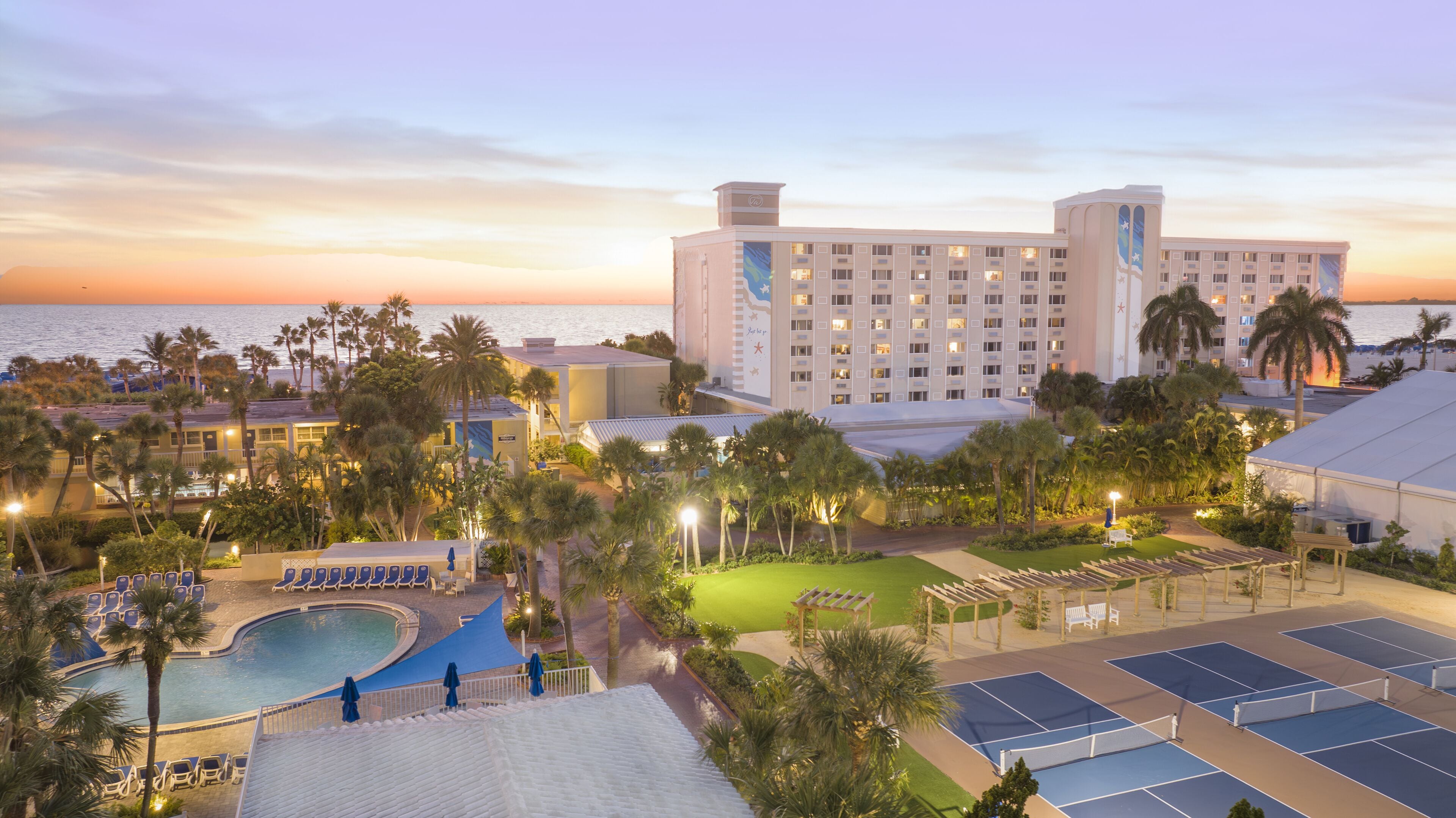 A hotel with a pool, tennis courts, and a view of the ocean at sunset.