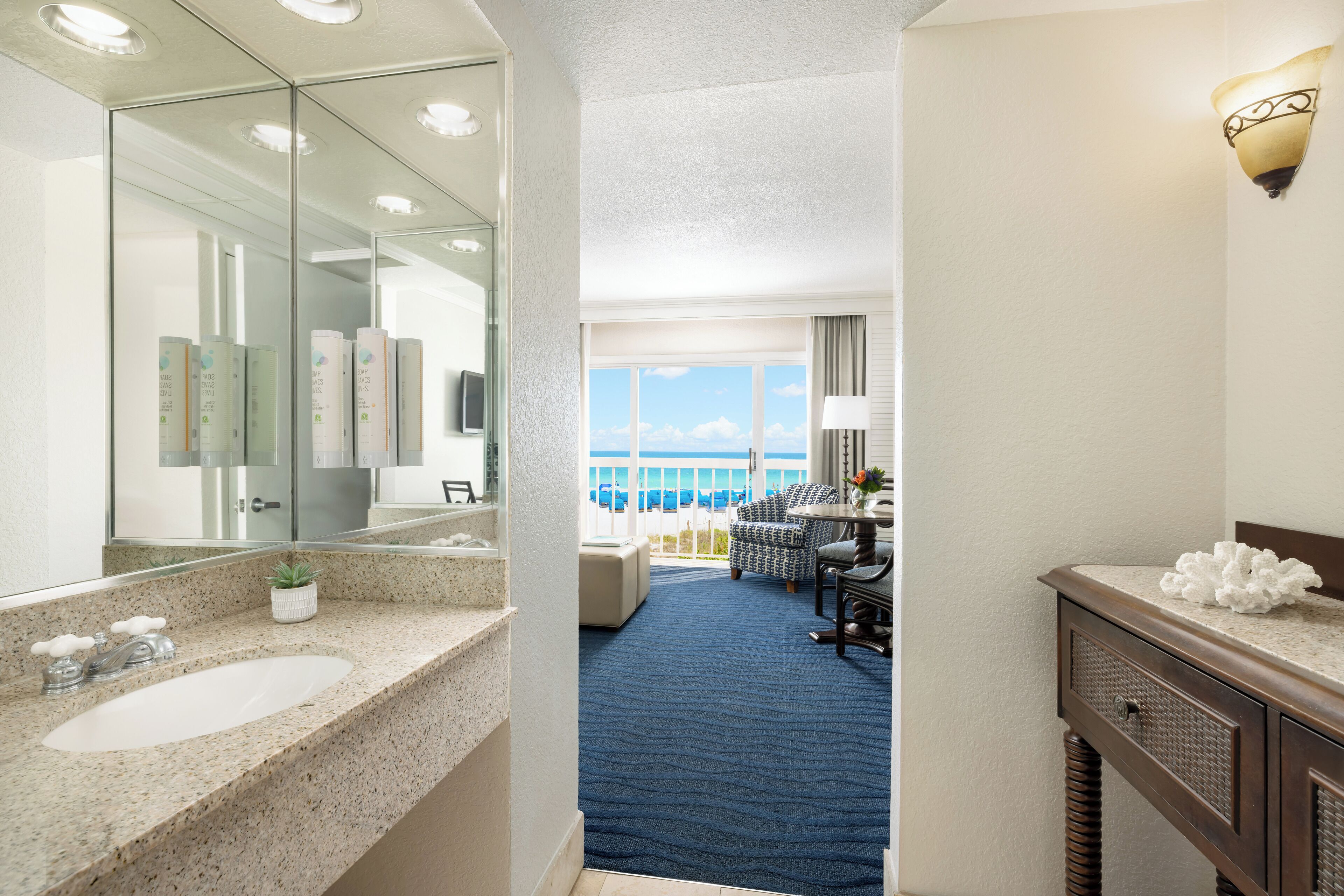 A hotel bathroom with a large mirror, a sink, and a vanity. There is a blue carpet leading to a balcony with a view of the ocean.