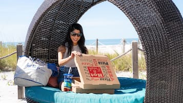 A person seated in a wicker chair with a beach view, holding a pizza box and a drink.