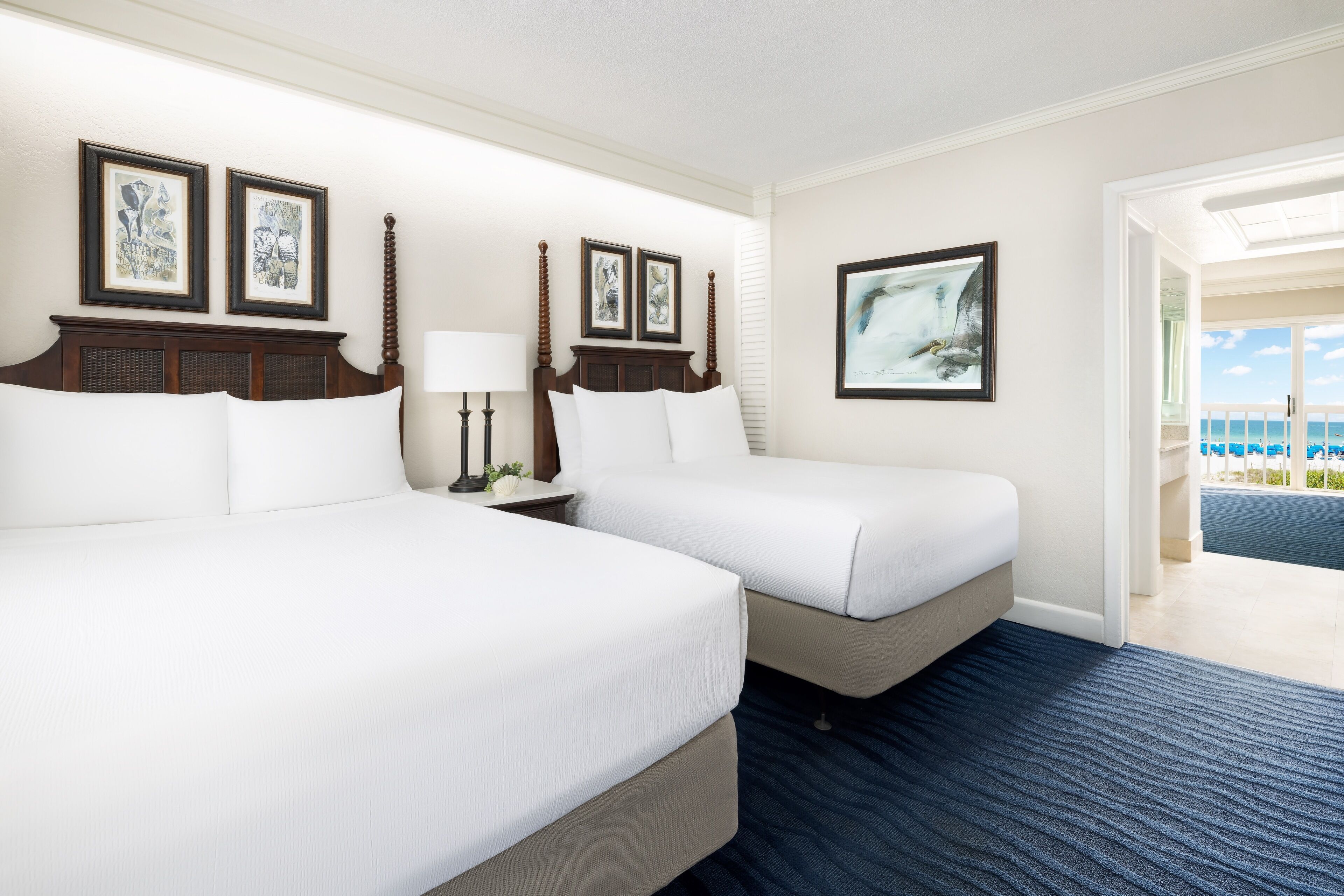 Habitación de hotel con dos camas, una mesita de noche, una lámpara y cuadros en las paredes.