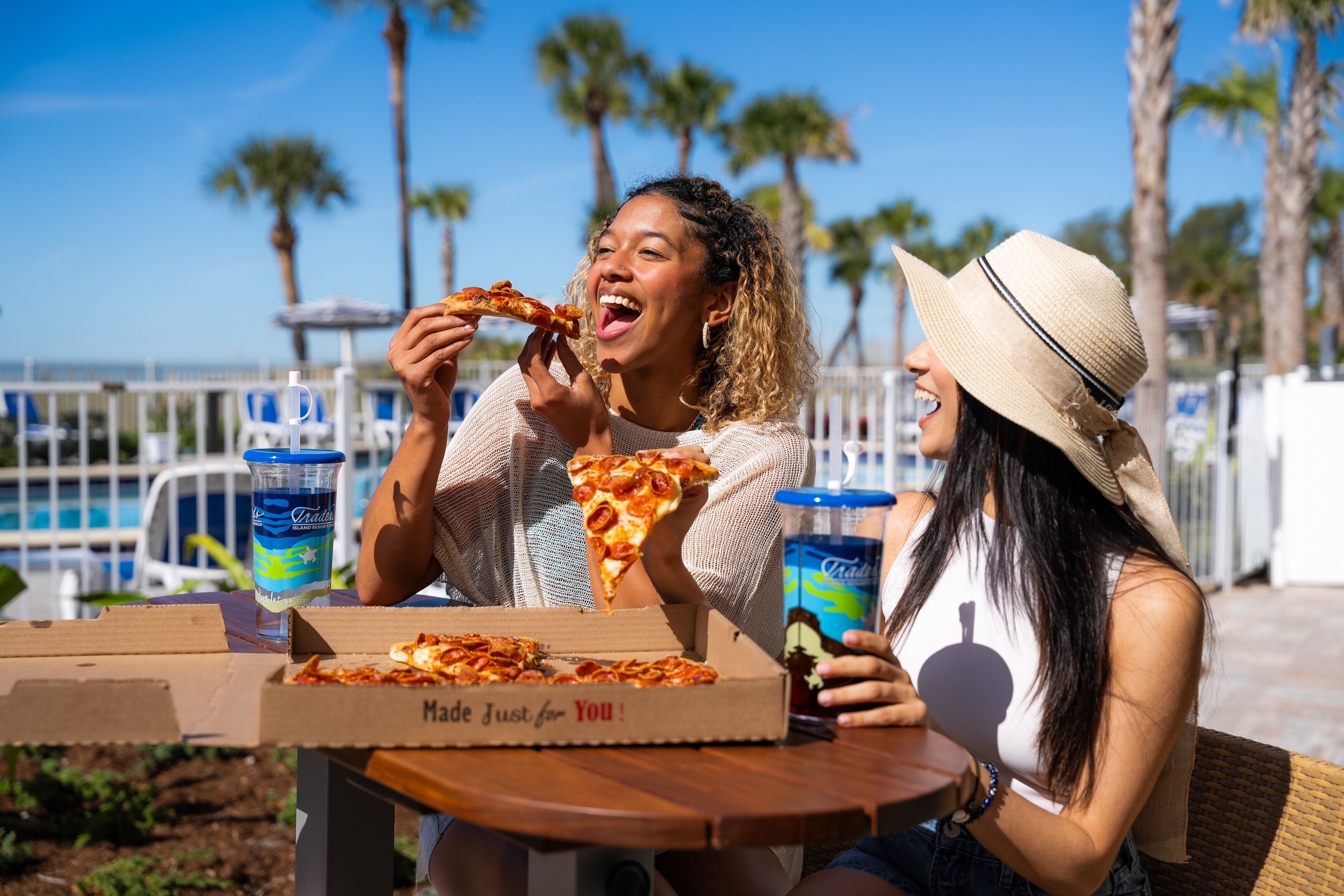 Two people sitting at a table with a pizza box and drinks.