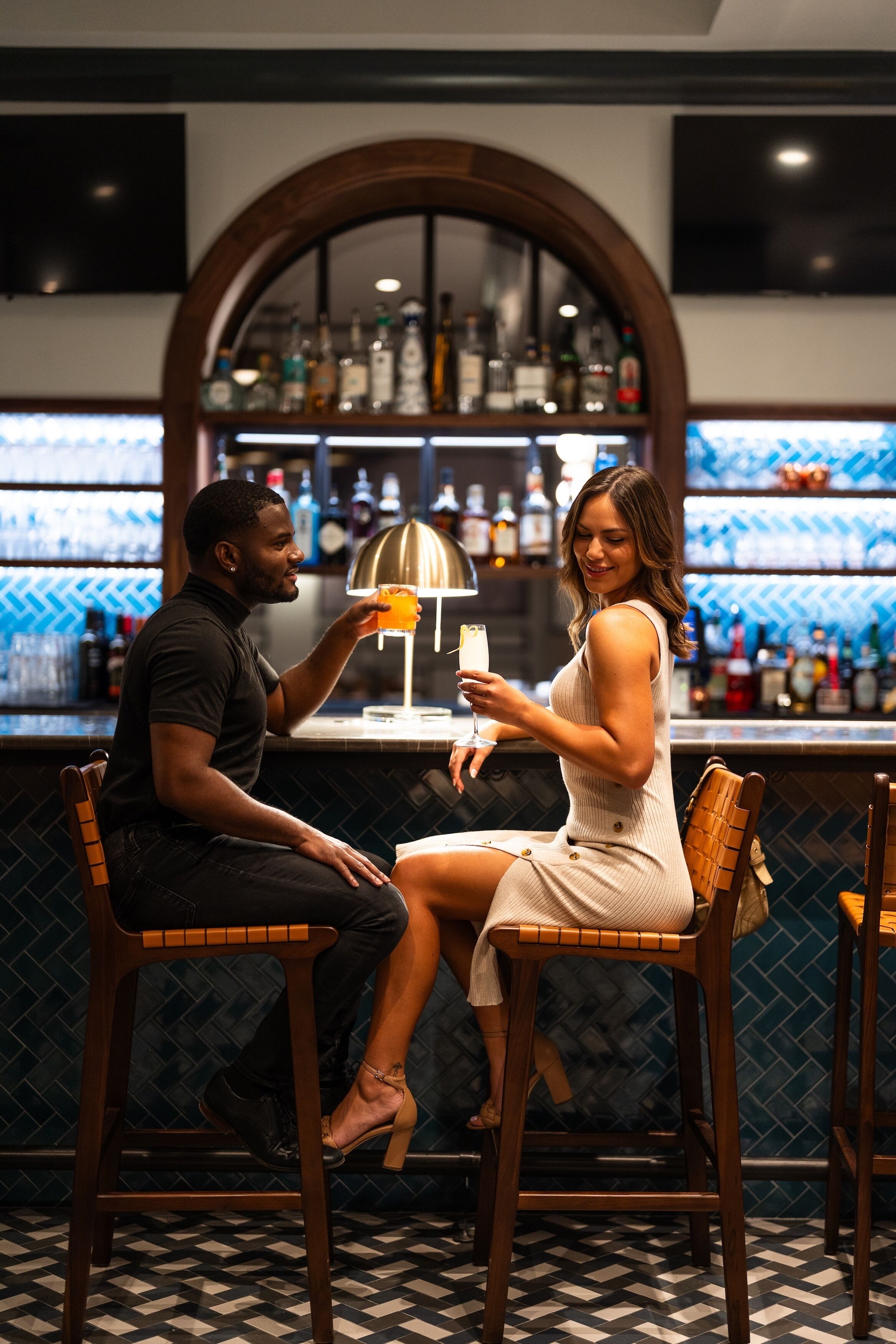 Una zona de bar con dos personas sentadas en la barra, un televisor colgado en la pared y una variedad de botellas en estanterías detrás de la barra.