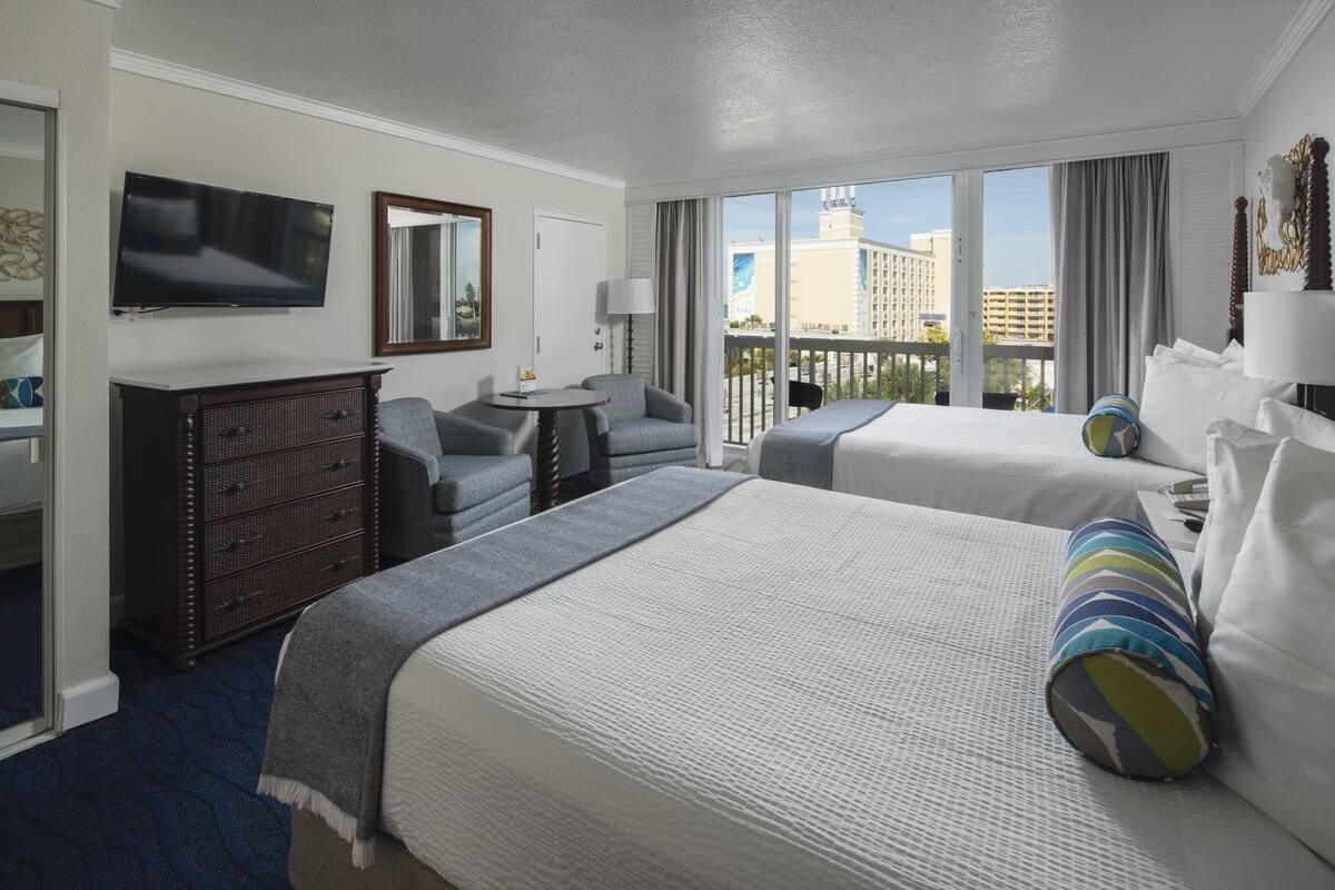 A hotel room with two beds, a TV, a dresser, and a balcony with a city view.
