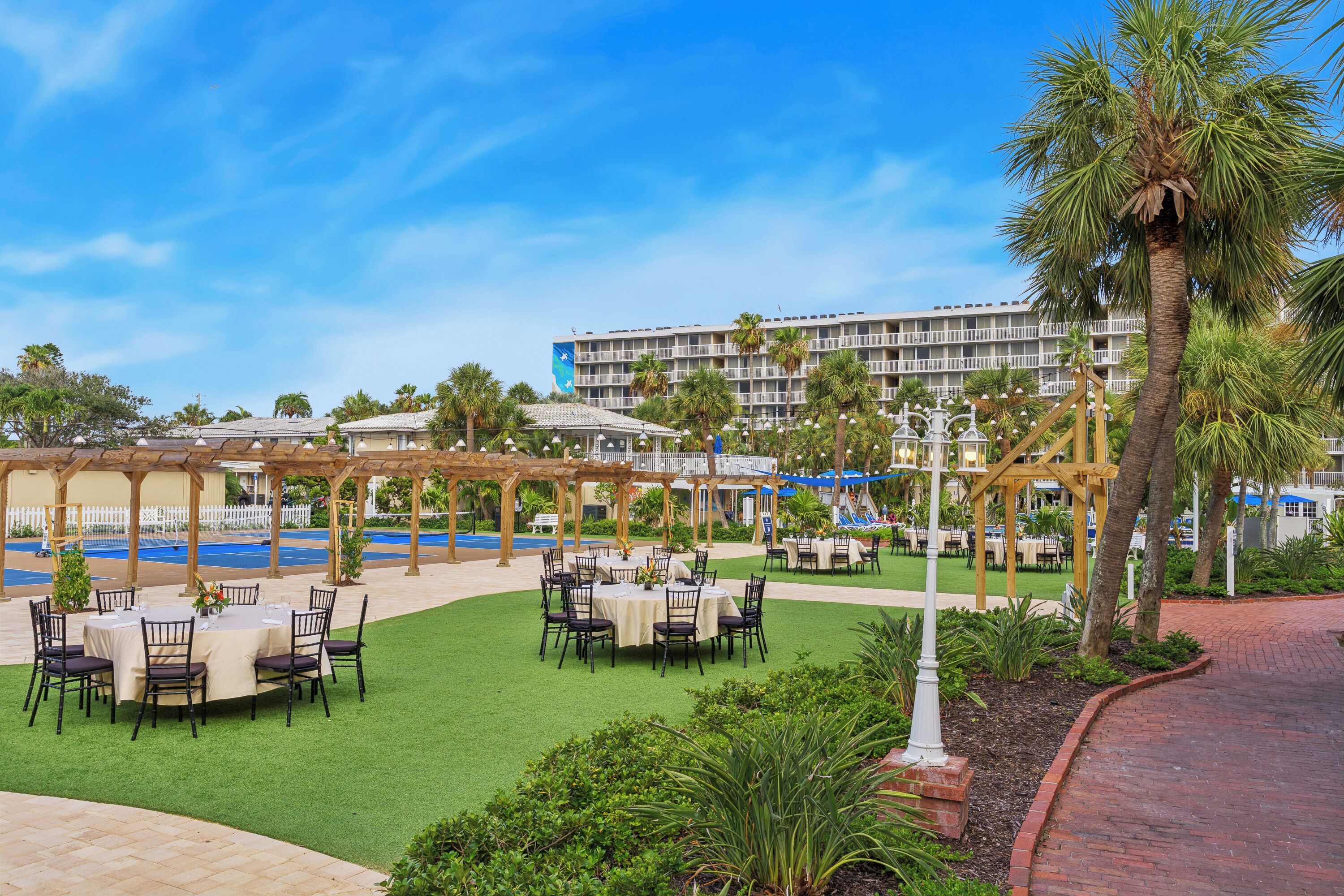 An outdoor dining area with tables and chairs, surrounded by palm trees and a pool.