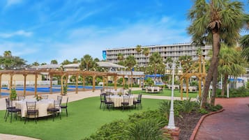 An outdoor dining area with tables and chairs, surrounded by palm trees and a pool.