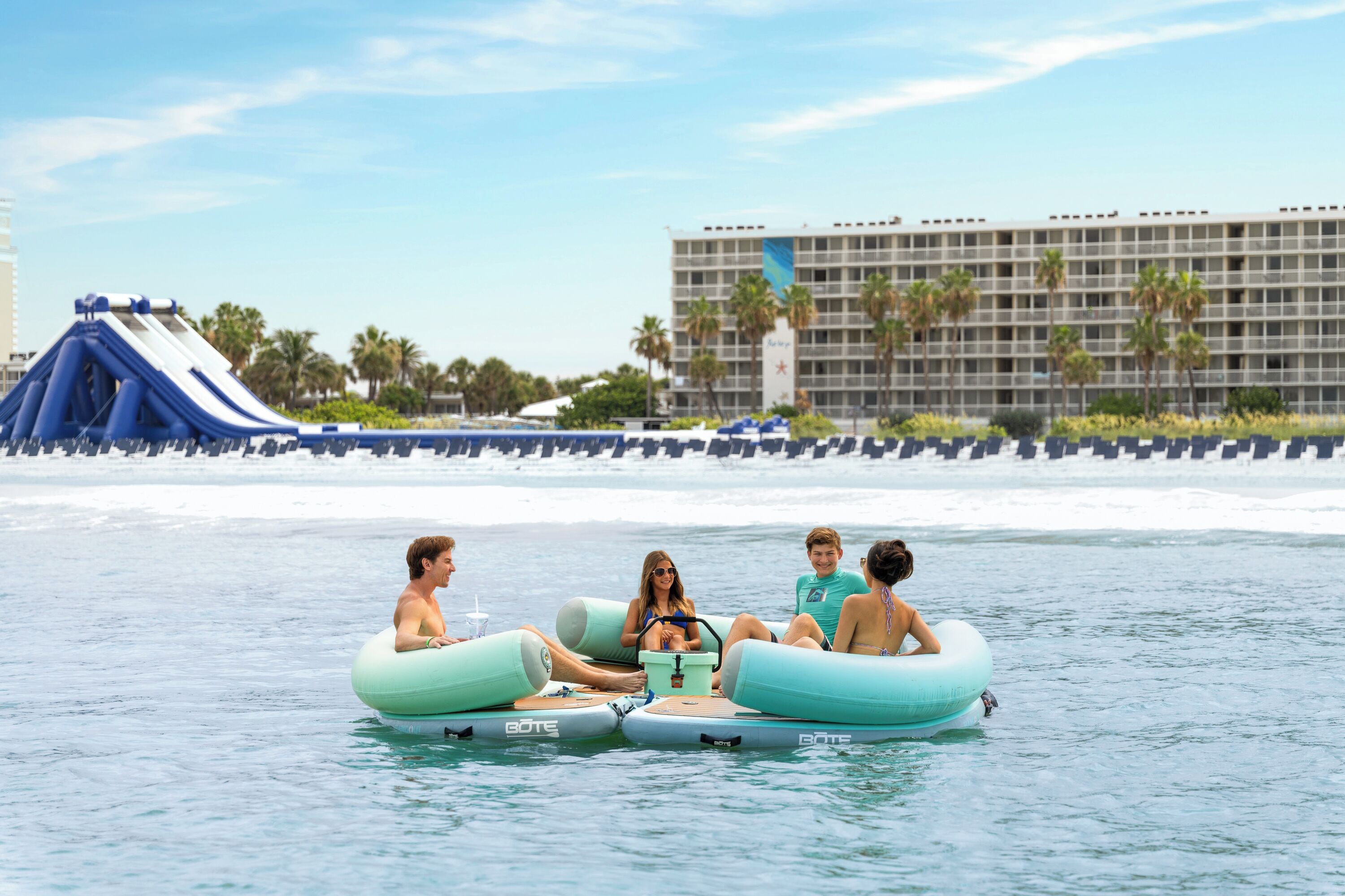 A group of people on inflatable rafts in a body of water with a building and palm trees in the background.