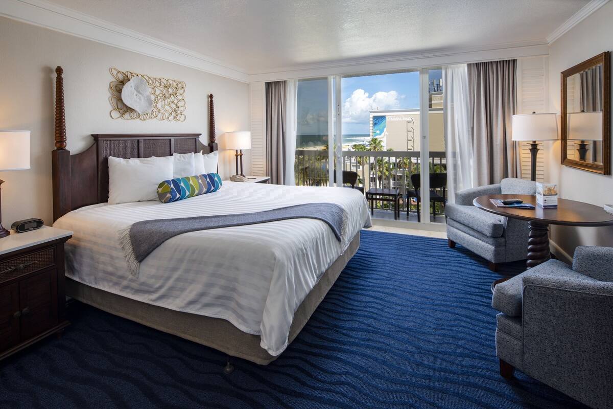 A hotel room with a large bed, a balcony with a view, a seating area, and a desk.
