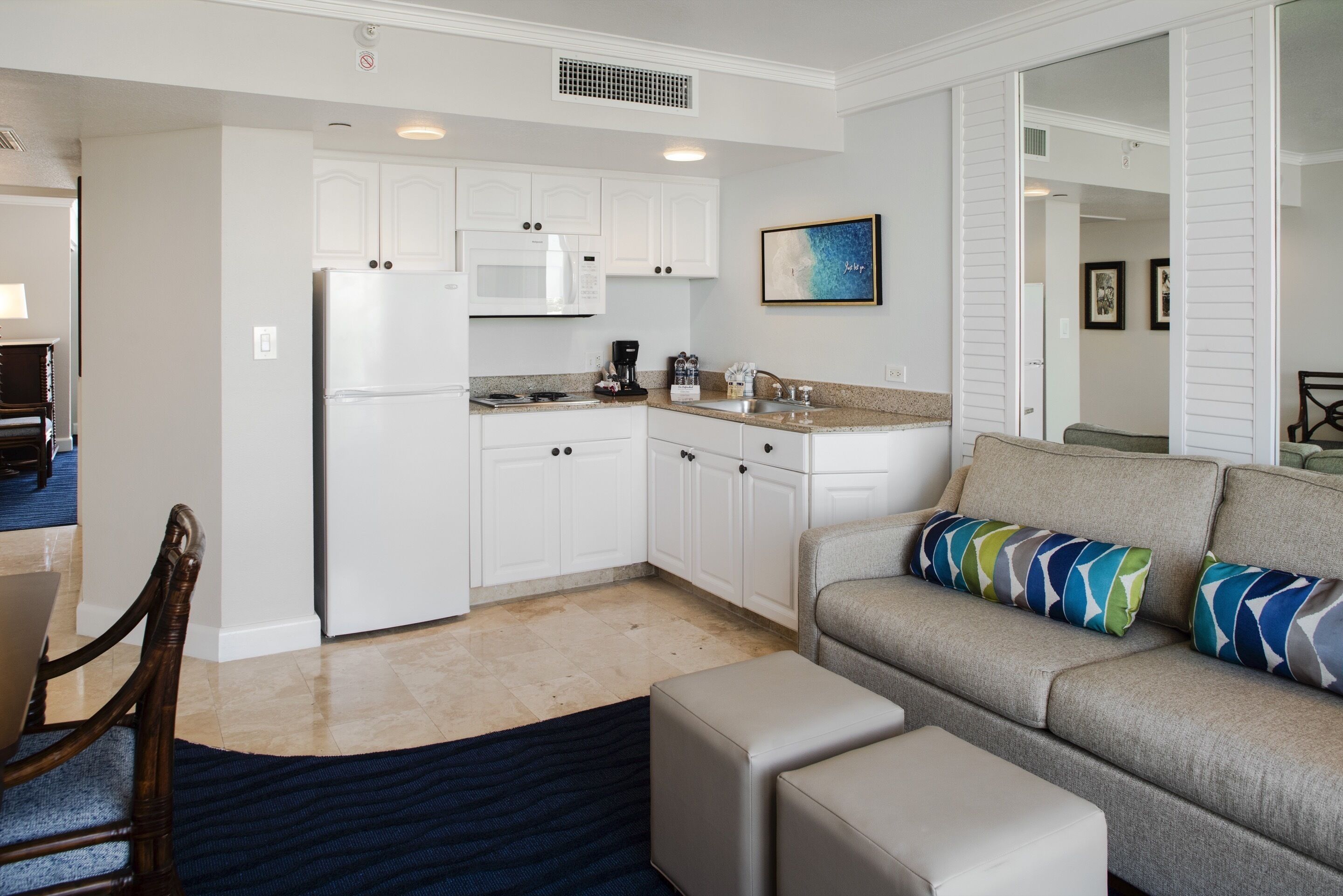 A modern kitchen with white cabinets, a microwave, and a coffee maker. There is a gray sofa with colorful pillows and a dark blue rug.