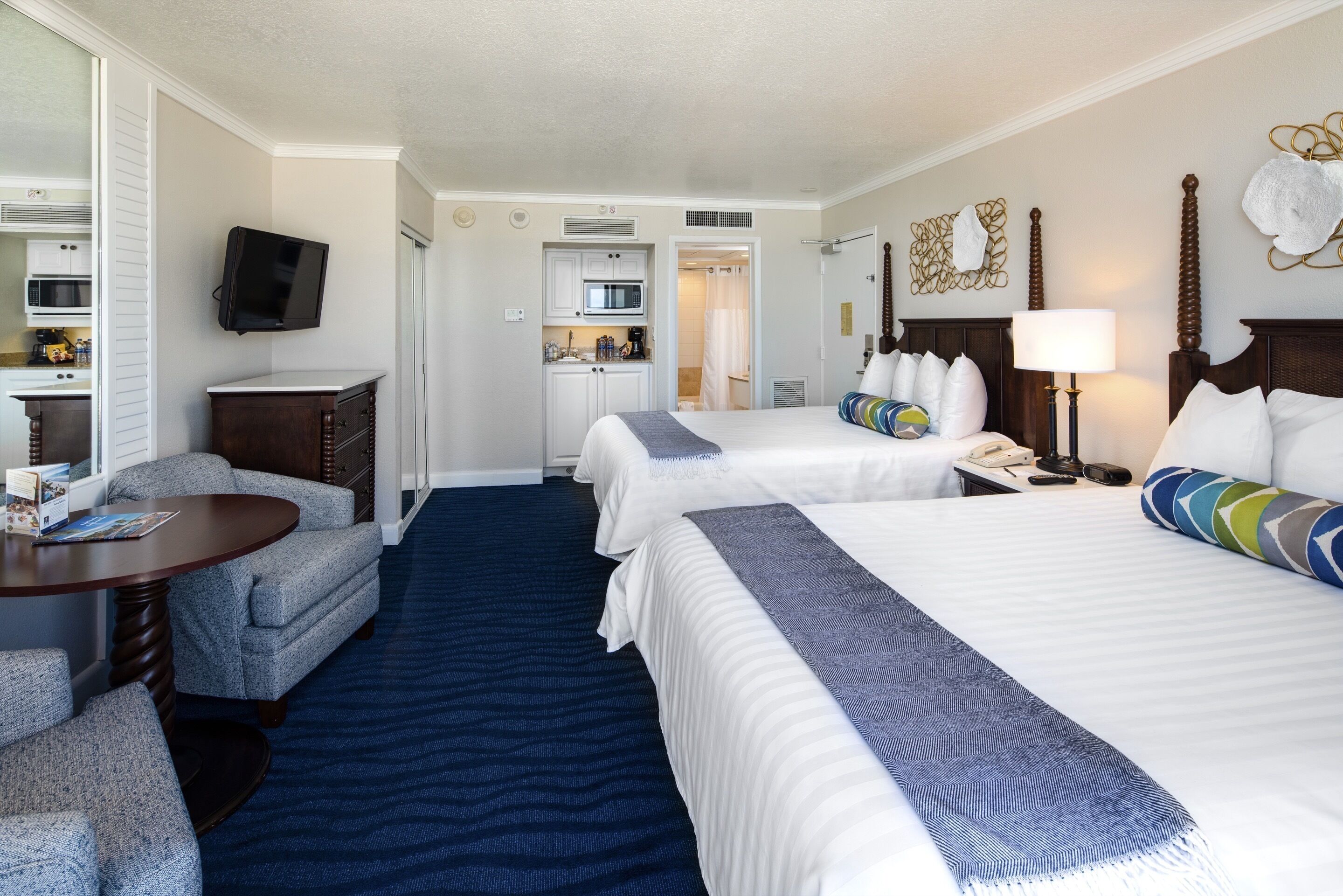 A hotel room with two beds, a desk, a chair, a TV, and a kitchenette.