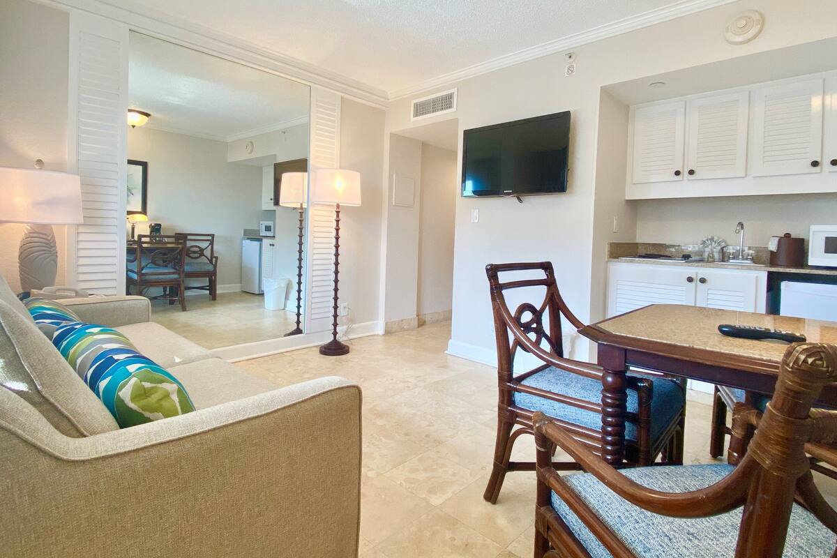 A living room with a sofa, a dining table with chairs, a lamp, and a TV mounted on the wall.