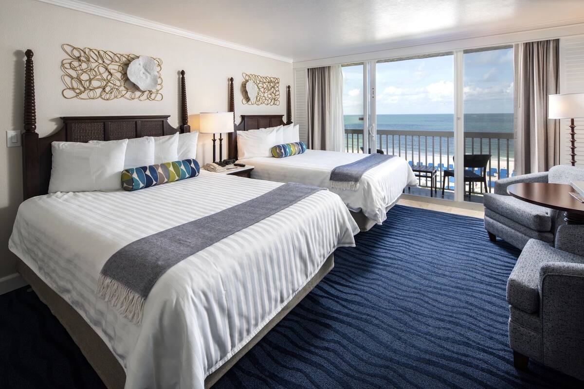 A hotel room with two beds, a balcony with a view of the ocean, and a seating area with a table and chairs.